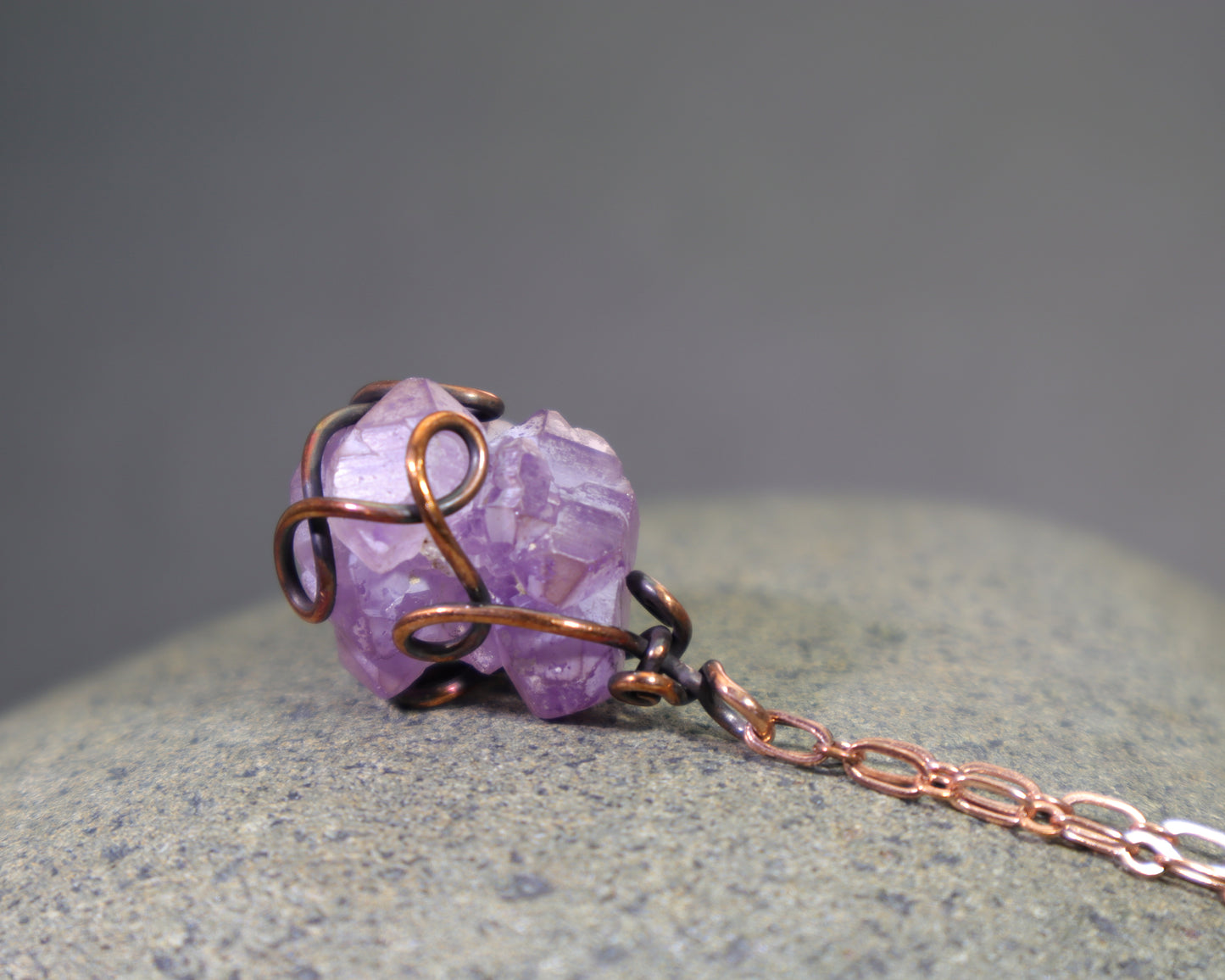Purple amethyst crystal pendant with copper wire wrapping on a gray rock