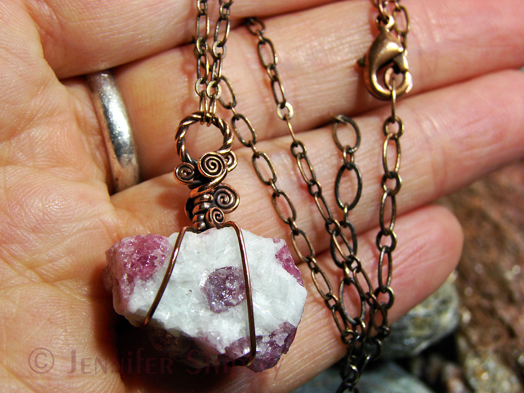 Raw pink tourmaline crystals in an albite matrix wire wrapped with copper on and adjustable chain, held in hand.