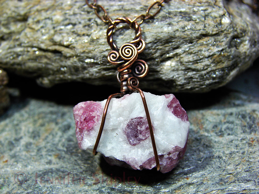 Raw pink tourmaline pendant necklace featuring natural rubellite crystals in white albite matrix wire wrapped in oxidized copper on a rock background