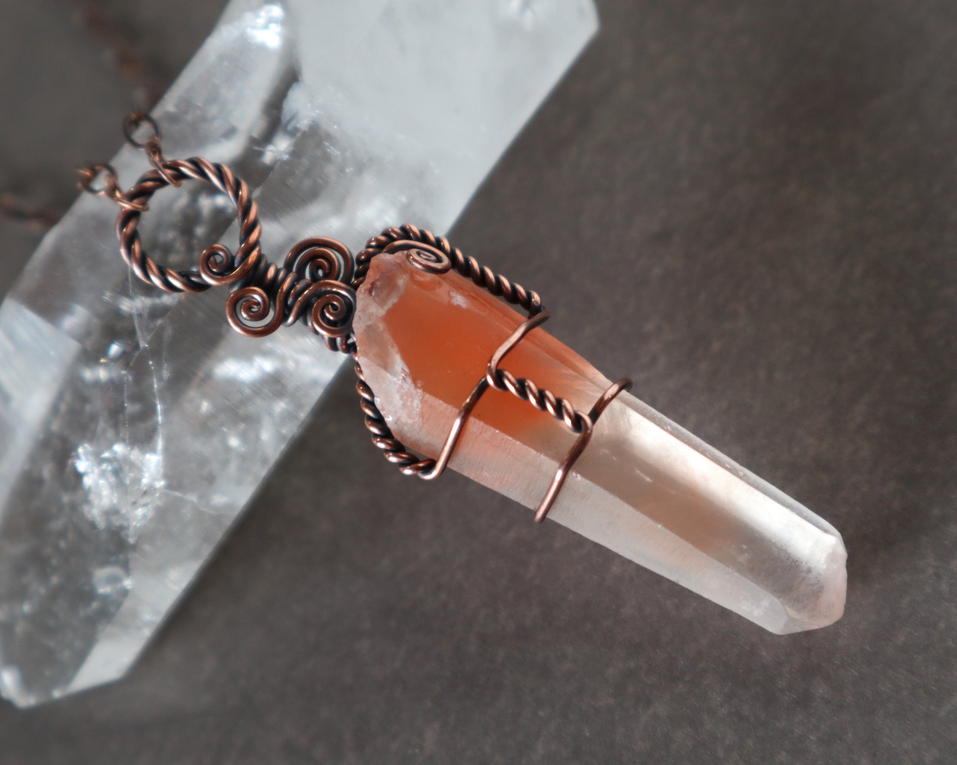 Red phantom quartz pendant necklace wire wrapped in oxidized copper shown on a gray surface leaning against a clear quartz crystal.