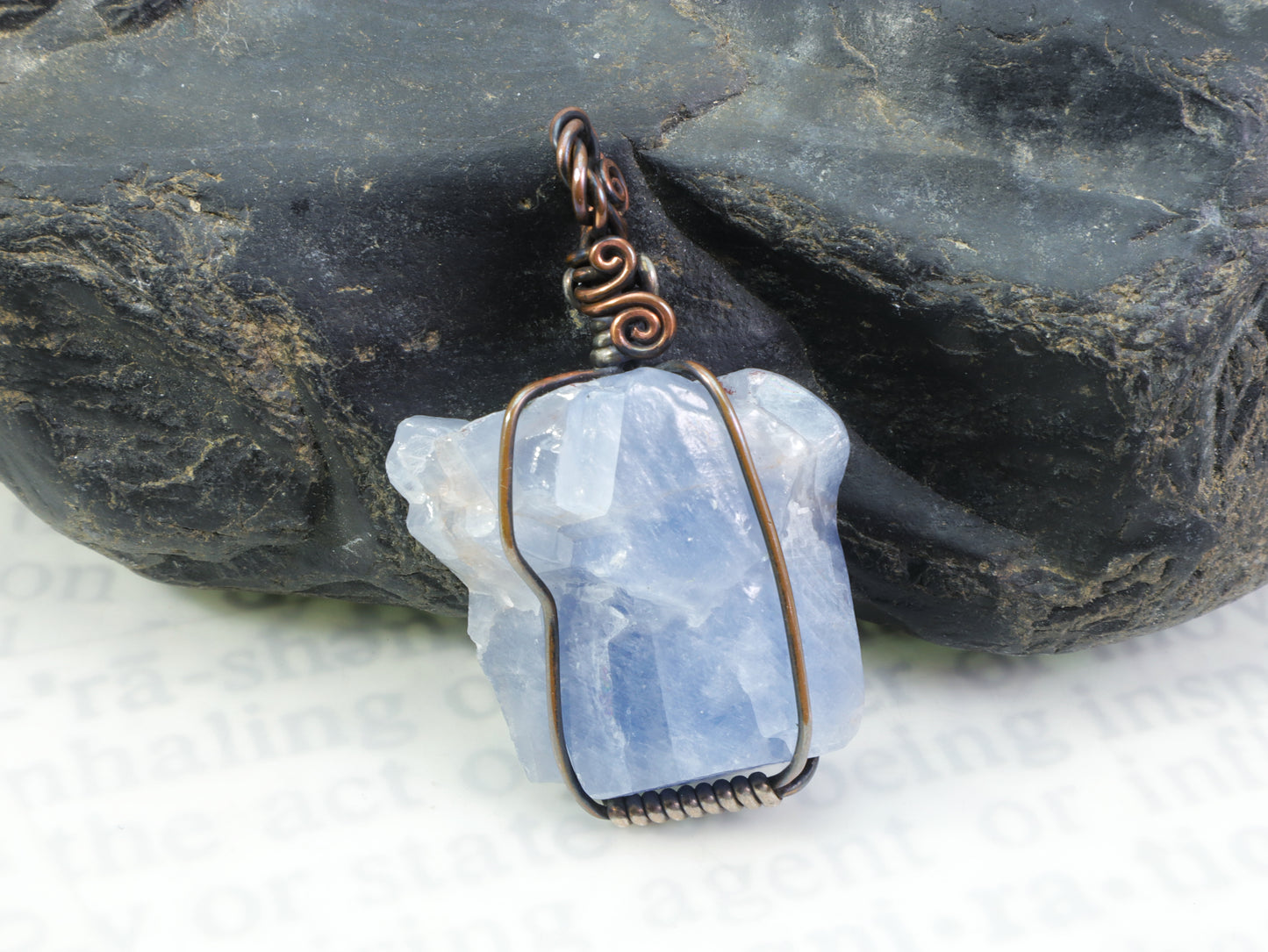 Blue calcite crystal pendant wire wrapped in copper and sterling silver in front of a black rock