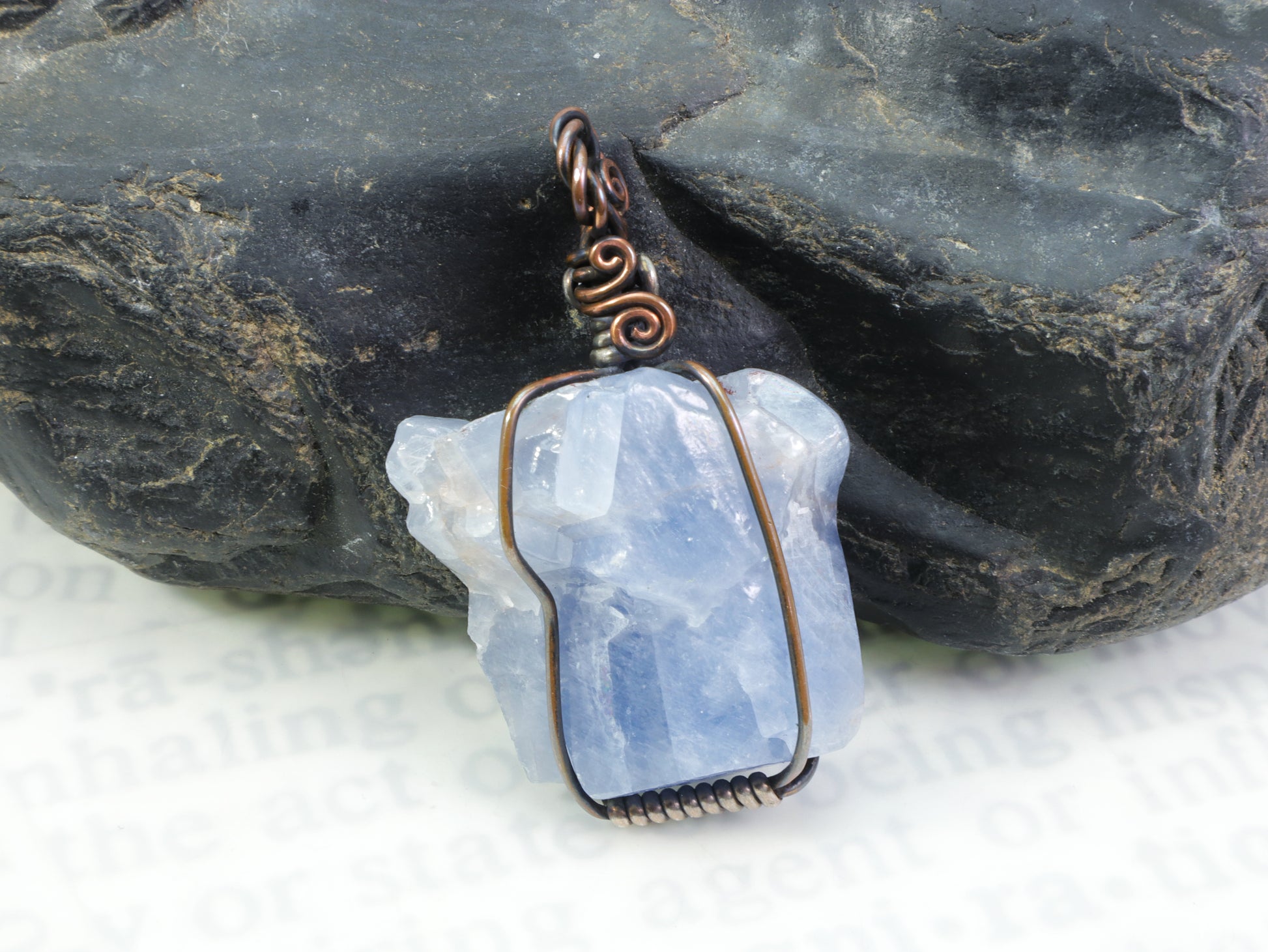 Blue calcite crystal pendant wire wrapped in copper and sterling silver in front of a black rock