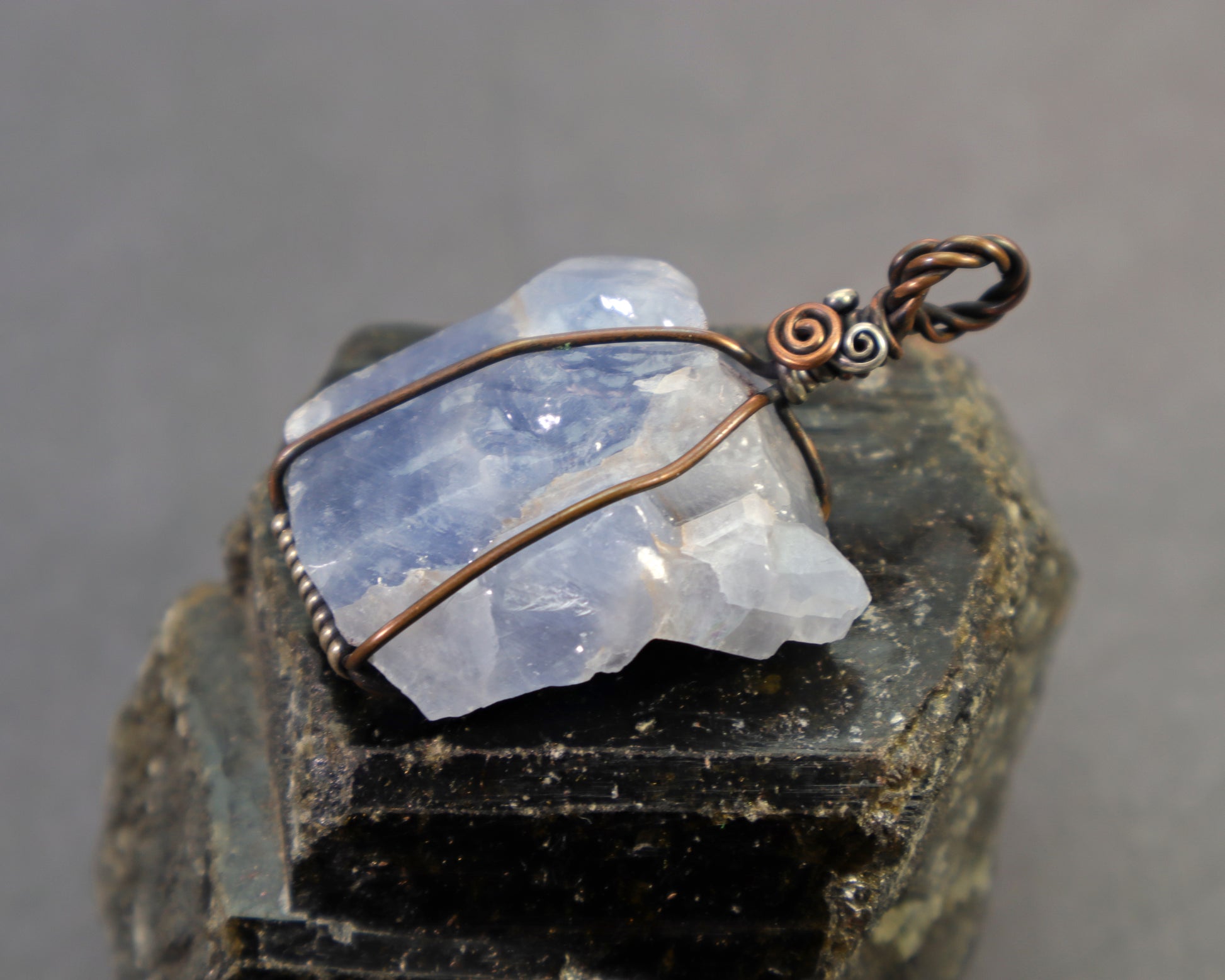 Raw blue calcite crystal pendant with art nouveau inspired wire wrapping sitting on to of a large black tourmaline crystal