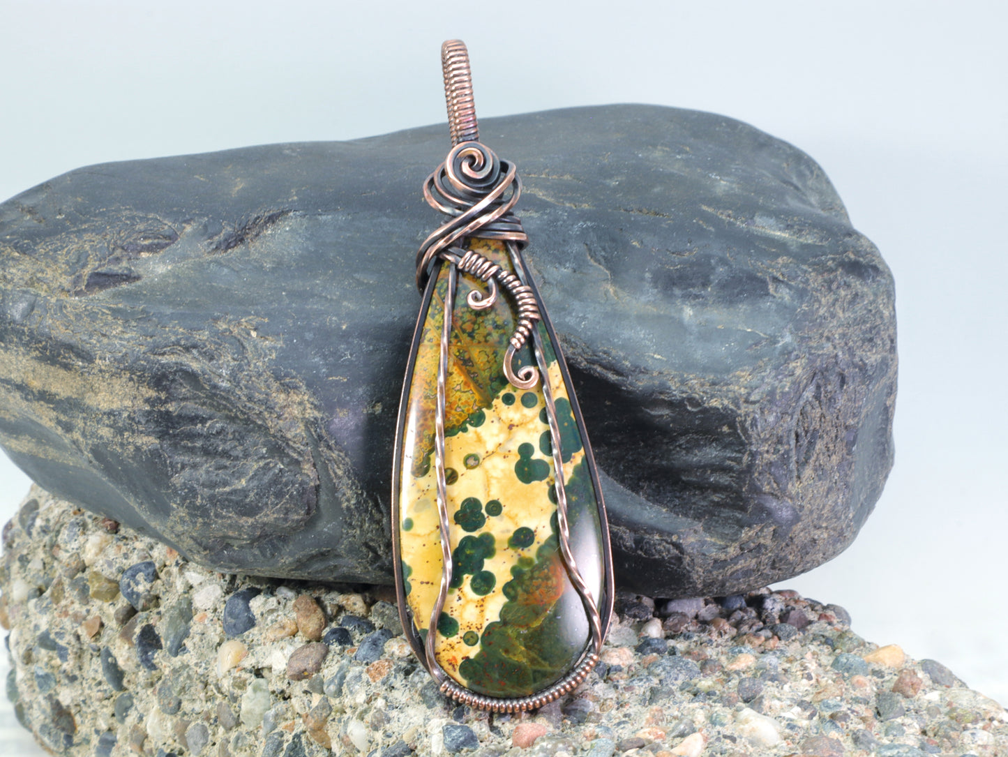Ocean Jasper pendant wire wrapped in oxidized copper sitting on a background of rocks