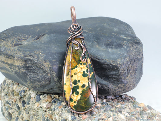 Ocean Jasper pendant wire wrapped in oxidized copper sitting on a background of rocks