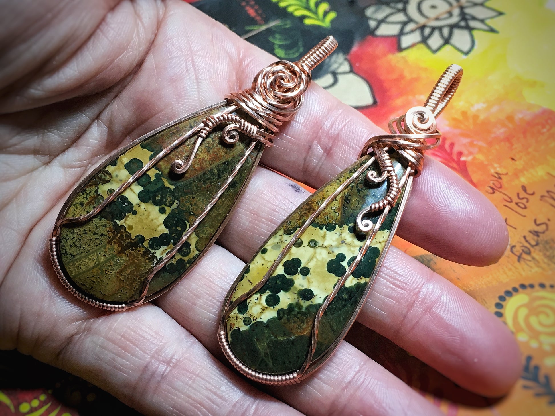 Two copper wrapped Ocean Jasper gemstones showcasing natural orb formations held in hand in front of one of the artist's paintings