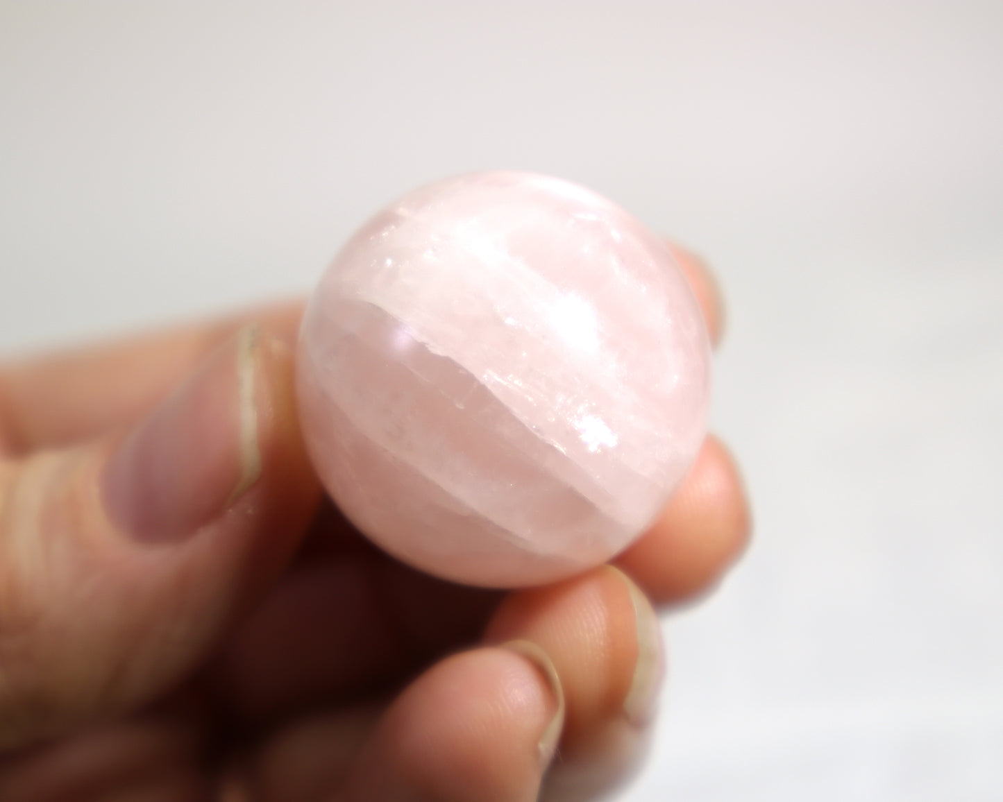 Natural pink quartz sphere with subtle white marbling held between fingers against a white background